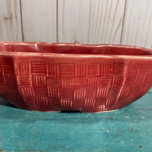 Puede incluir: Una jardinera de cerámica roja con textura de cesta tejida. La jardinera rectangular tiene una forma ligeramente curvada y es adecuada para uso en interiores o exteriores. La jardinera es de color rojo liso.