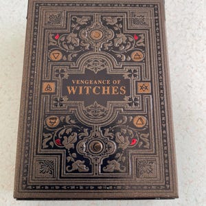 May include: A decorative box with the text "Vengeance of Witches" in gold lettering. The box has a dark brown background with ornate, embossed floral and geometric designs in a lighter brown color. Red accents are visible.