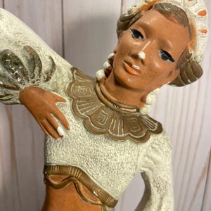 Puede incluir: Una figurita de cerámica de una mujer con un vestido blanco y dorado y una cubierta de cabeza blanca. La mujer está sosteniendo su brazo hacia arriba y tiene una tez morena.