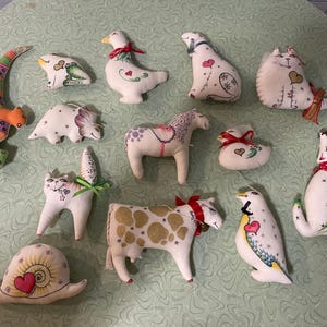 Könnte beinhalten: Eine Sammlung handgefertigter Stofftierornamente, darunter eine Schnecke, eine Katze, eine Kuh, ein Pferd, ein Pinguin und ein Hund. Jedes Ornament ist weiß mit farbenfrohen Details und Akzenten wie Bändern und Herzen.