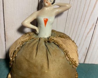 May include: A vintage porcelain pincushion with a ballerina figurine. The ballerina is wearing a white dress with a red heart and a blue headband. The pincushion is covered in a brown fabric with gold trim.