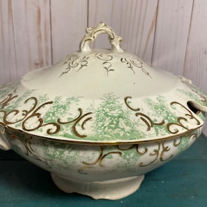 May include: A large white porcelain serving bowl with a lid. The bowl and lid are decorated with a green and gold floral pattern. The bowl has a round base and a wide, rounded rim. The lid has a handle on top.