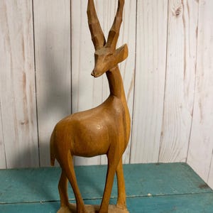 Könnte beinhalten: Eine Holzskulptur eines braunen Antilopen mit langen, gebogenen Hörnern. Die Skulptur steht auf einem Holzsockel.