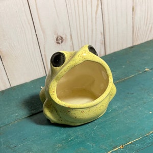 May include: A yellow ceramic frog with black eyes, speckled with brown spots, is shaped like a dish with an open mouth. The frog is a decorative dish.