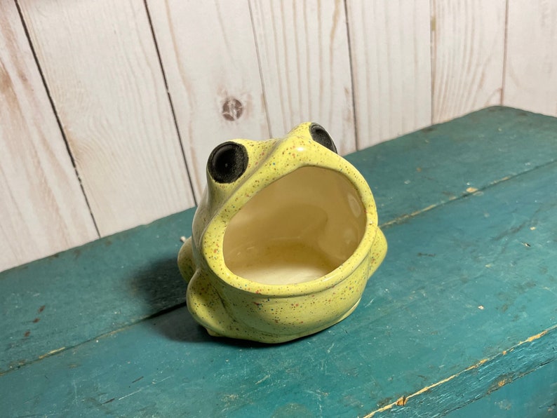 May include: A yellow ceramic frog with black eyes, speckled with brown spots, is shaped like a dish with an open mouth. The frog is a decorative dish.