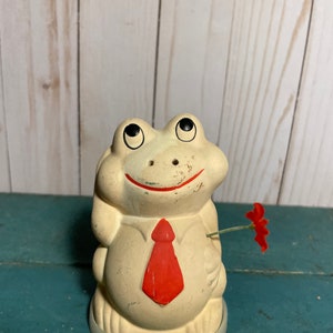 May include: A white ceramic frog figurine wearing a red tie and holding a red flower. The frog has black eyes and a smiling mouth.