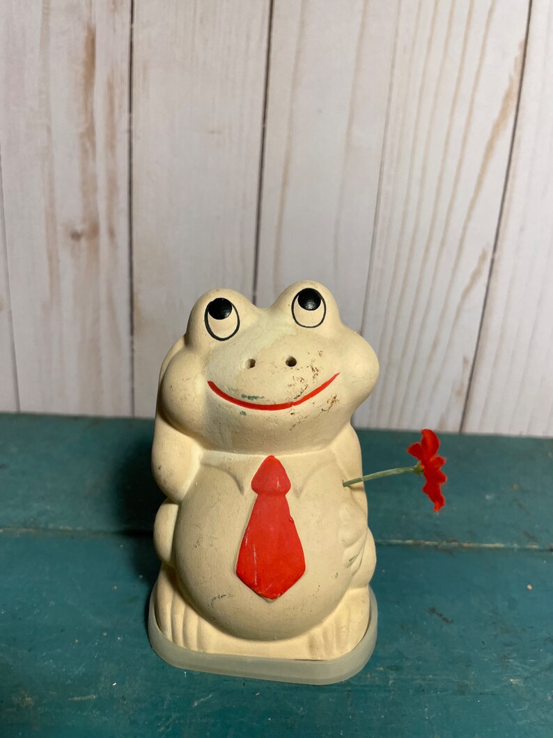 May include: A white ceramic frog figurine wearing a red tie and holding a red flower. The frog has black eyes and a smiling mouth.