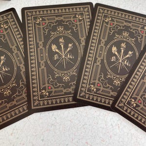 May include: Four playing cards with a dark brown background and gold decorative borders. Each card features a central emblem of crossed torches and spears within a circle, with red accents. The cards are arranged on a light surface.