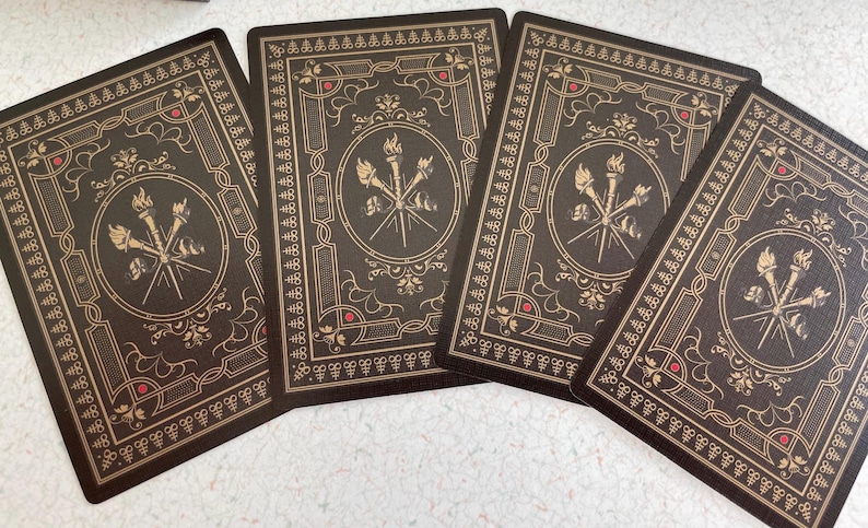 May include: Four playing cards with a dark brown background and gold decorative borders. Each card features a central emblem of crossed torches and spears within a circle, with red accents. The cards are arranged on a light surface.