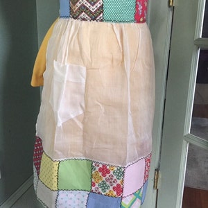 May include: A vintage-style apron with a patchwork design. The apron features a cream-colored body with a colorful patchwork waistband and hem. The patchwork includes various patterns and colors, including floral, polka dots, and geometric shapes.