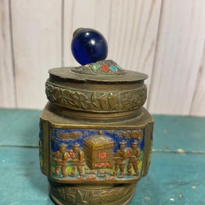 Könnte beinhalten: Eine kleine, runde, messingfarbene Schmuckschatulle mit einem blauen Glasknopf auf dem Deckel. Die Schachtel ist mit buntem Emaille und aufwendigen Schnitzereien verziert. Die Schachtel zeigt eine Szene von Menschen, die eine Sänfte tragen.