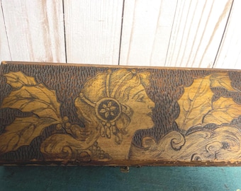 May include: A wooden box with a carved design of a woman's face and leaves. The box is brown and has a textured surface.