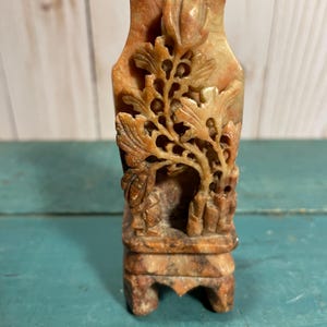 Könnte beinhalten: Eine kleine, aufwendig geschnitzte Steinvase in Beige- und Brauntönen. Die Vase hat ein detailliertes Blumendesign mit rechteckigem Sockel. Die Schnitzerei ist auf der Vorderseite der Vase sichtbar. Die Vase ist etwa 10 cm hoch.