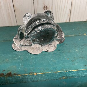 May include: A clear glass frog figurine with a detailed design. The frog is sitting on a textured base.