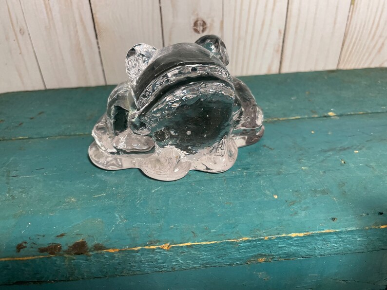 May include: A clear glass frog figurine with a detailed design. The frog is sitting on a textured base.