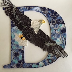 Può includere: Un'opera d'arte in quilling di carta che rappresenta un'aquila calva in volo, formando la lettera D. L'aquila è realizzata con carta nera, bianca e gialla, e lo sfondo è realizzato con carta blu e bianca.