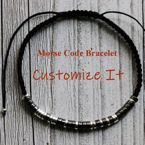 May include: A black cord bracelet with silver beads that spell out a message in Morse code. The bracelet is adjustable and has a sliding knot. The text on the image reads "Morse Code Bracelet Customize It".