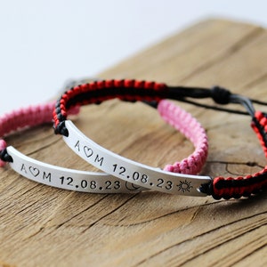 May include: Two matching bracelets with black and red, and black and pink woven cords. Each bracelet has a silver bar with the inscription "A&hearts;M 12.08.23" and symbols of a moon and sun.