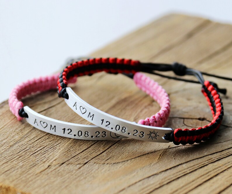 May include: Two matching bracelets with black and red, and black and pink woven cords. Each bracelet has a silver bar with the inscription "A&hearts;M 12.08.23" and symbols of a moon and sun.