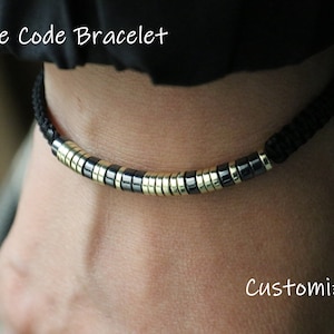 May include: A black cord bracelet with gold and black beads. The bracelet is on a person's wrist. The text "Morse Code Bracelet" and "Customize It" is visible in the image.
