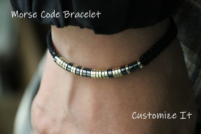 May include: A black cord bracelet with gold and black beads. The bracelet is on a person's wrist. The text "Morse Code Bracelet" and "Customize It" is visible in the image.