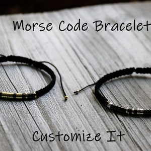 May include: Two black cord bracelets with gold and silver beads. The bracelets are on a wooden surface. The text "Morse Code Bracelet" and "Customize It" are visible.