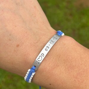May include: A blue and white braided bracelet with a silver metal charm that says "JCD 03.12.24" with a heart symbol.