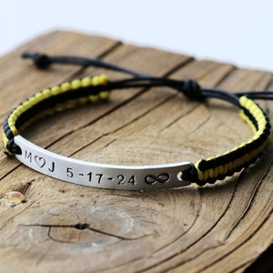 May include: A black and yellow braided bracelet with a silver bar charm engraved with the initials "M&hearts;J" and the date "5-17-24" followed by an infinity symbol.