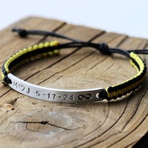 May include: A black and yellow braided bracelet with a silver bar charm engraved with the initials "M&hearts;J" and the date "5-17-24" followed by an infinity symbol.