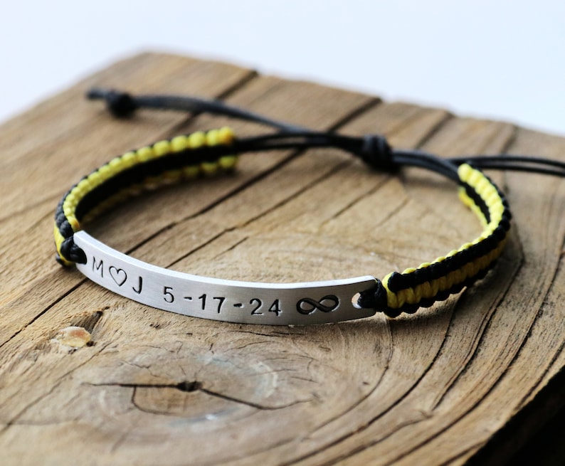 May include: A black and yellow braided bracelet with a silver bar charm engraved with the initials "M&hearts;J" and the date "5-17-24" followed by an infinity symbol.