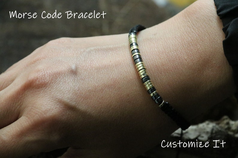 May include: A black cord bracelet with gold and black beads. The bracelet is on a person's wrist. The text "Morse Code Bracelet" and "Customize It" is visible in the image.