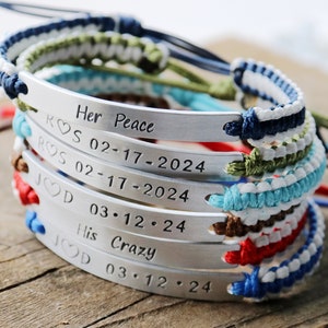 May include: A stack of five bracelets with silver metal tags engraved with the text "Her Peace ROS 02-17-2024 ROS 02-17-2024 JOD 03.12.24 His Crazy JOD 03.12.24". The bracelets are made of colorful braided cords.