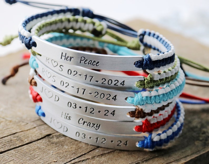 May include: A stack of five bracelets with silver metal tags engraved with the text "Her Peace ROS 02-17-2024 ROS 02-17-2024 JOD 03.12.24 His Crazy JOD 03.12.24". The bracelets are made of colorful braided cords.
