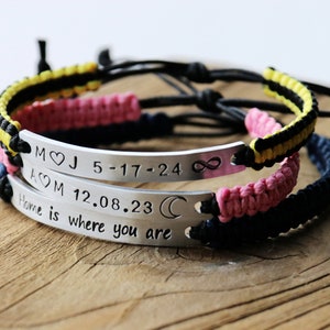 May include: Two silver bracelets with black, pink, and yellow braided cords. The bracelets have engraved metal plates with the text "M&hearts;J 5-17-24 &infin;" and "A&hearts;M 12.08.23 ☾ Home is where you are".