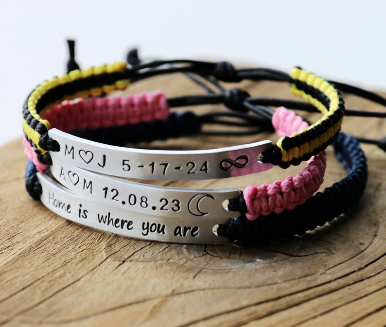 May include: Two silver bracelets with black, pink, and yellow braided cords. The bracelets have engraved metal plates with the text "M&hearts;J 5-17-24 &infin;" and "A&hearts;M 12.08.23 ☾ Home is where you are".