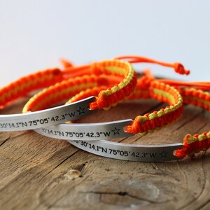May include: Several handmade bracelets with orange and yellow woven bands and silver bars. The bars are engraved with coordinates and a star or moon symbol. The coordinates read: 38&deg;30'14.1"N 75&deg;05'42.3"W.