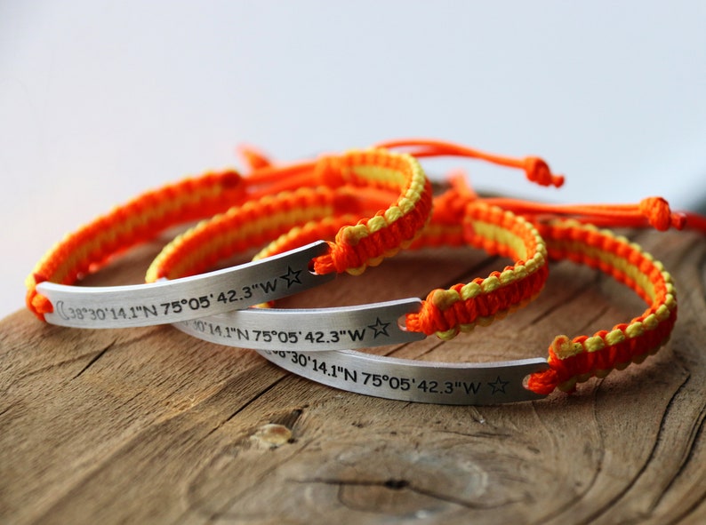 May include: Several handmade bracelets with orange and yellow woven bands and silver bars. The bars are engraved with coordinates and a star or moon symbol. The coordinates read: 38&deg;30'14.1"N 75&deg;05'42.3"W.