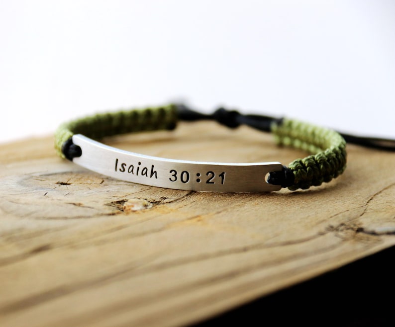 May include: A green and black braided bracelet with a silver metal tag that reads "Isaiah 30:21".