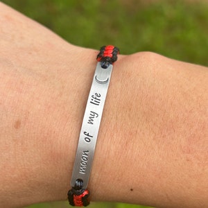 May include: A silver bracelet with a black and red braided cord. The bracelet has a silver tag with the words "moon of my life" engraved on it.