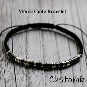 May include: A black and silver beaded bracelet with a black cord. The bracelet is adjustable and has a sliding knot. The bracelet is on a wooden surface. "Morse Code Bracelet" and "Customize It" are written on the surface.