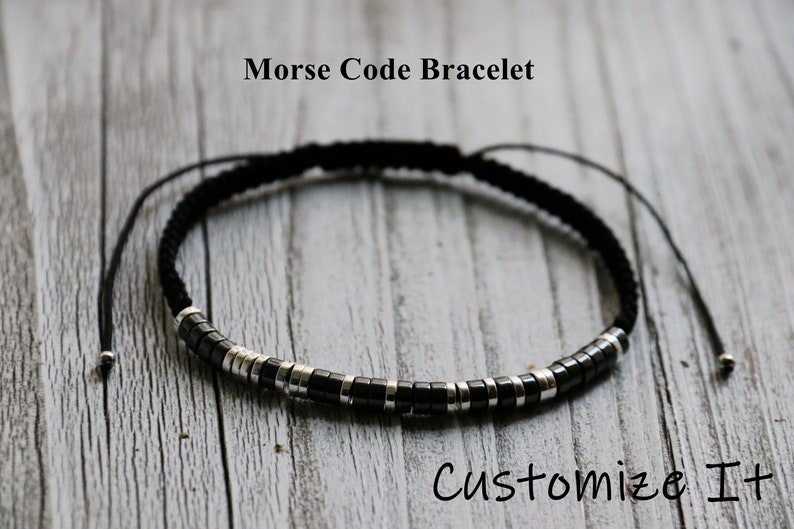 May include: A black and silver beaded bracelet with a black cord. The bracelet is adjustable and has a sliding knot. The bracelet is on a wooden surface. "Morse Code Bracelet" and "Customize It" are written on the surface.