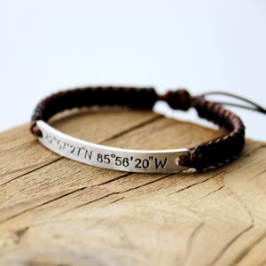 May include: A brown leather cord bracelet with a silver bar charm engraved with the coordinates "42°57'27"N 85°56'20"W".