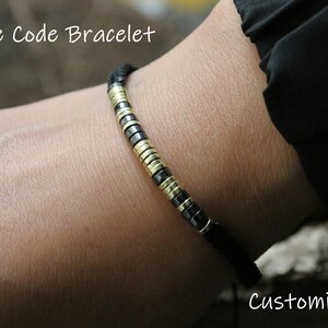 May include: A black cord bracelet with gold and black beads. The bracelet is on a person's wrist. The text "Morse Code Bracelet" is at the top of the image and the text "Customize It" is at the bottom of the image.