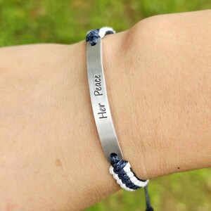 May include: A white and navy blue braided bracelet with a silver bar charm that says "Her Peace".
