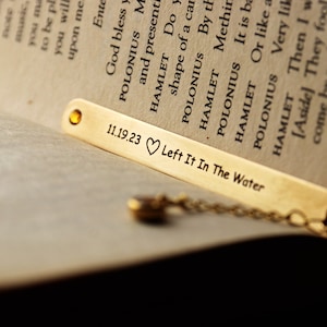 May include: A gold bookmark with a chain attached. The bookmark is engraved with the date "11.19.23", a heart symbol, and the text "Left It In The Water".