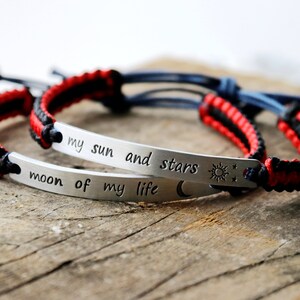 May include: Two red and black braided bracelets with silver metal charms. The charms have the text "my sun and stars" and "moon of my life" engraved on them.