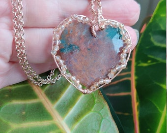May include: A heart-shaped pendant necklace with a wire-wrapped, multi-colored stone. The stone has shades of brown, green, and orange. The pendant is surrounded by small, clear beads.