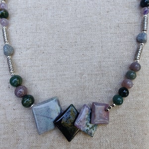 Puede incluir: Un collar de piedras preciosas con una variedad de piedras naturales. El collar incluye piedras cuadradas y en forma de diamante en tonos grises, negros y morados, intercaladas con cuentas redondas verdes y moradas. Acentos metálicos plateados conectan las piedras.