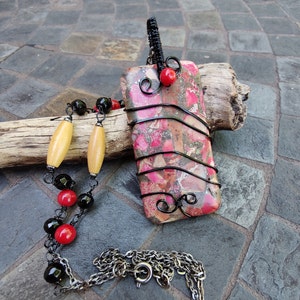 Pode incluir: Um colar com pingente retangular com uma pedra rosa, vermelha e marrom envolta em arame preto. O colar apresenta uma corrente com contas pretas, vermelhas e amarelas. O pingente tem aproximadamente 5 cm de comprimento.
