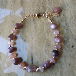 May include: A gold bracelet with a variety of light pink, purple, and gray gemstone beads. The bracelet has a gold clasp and a small gold charm.
